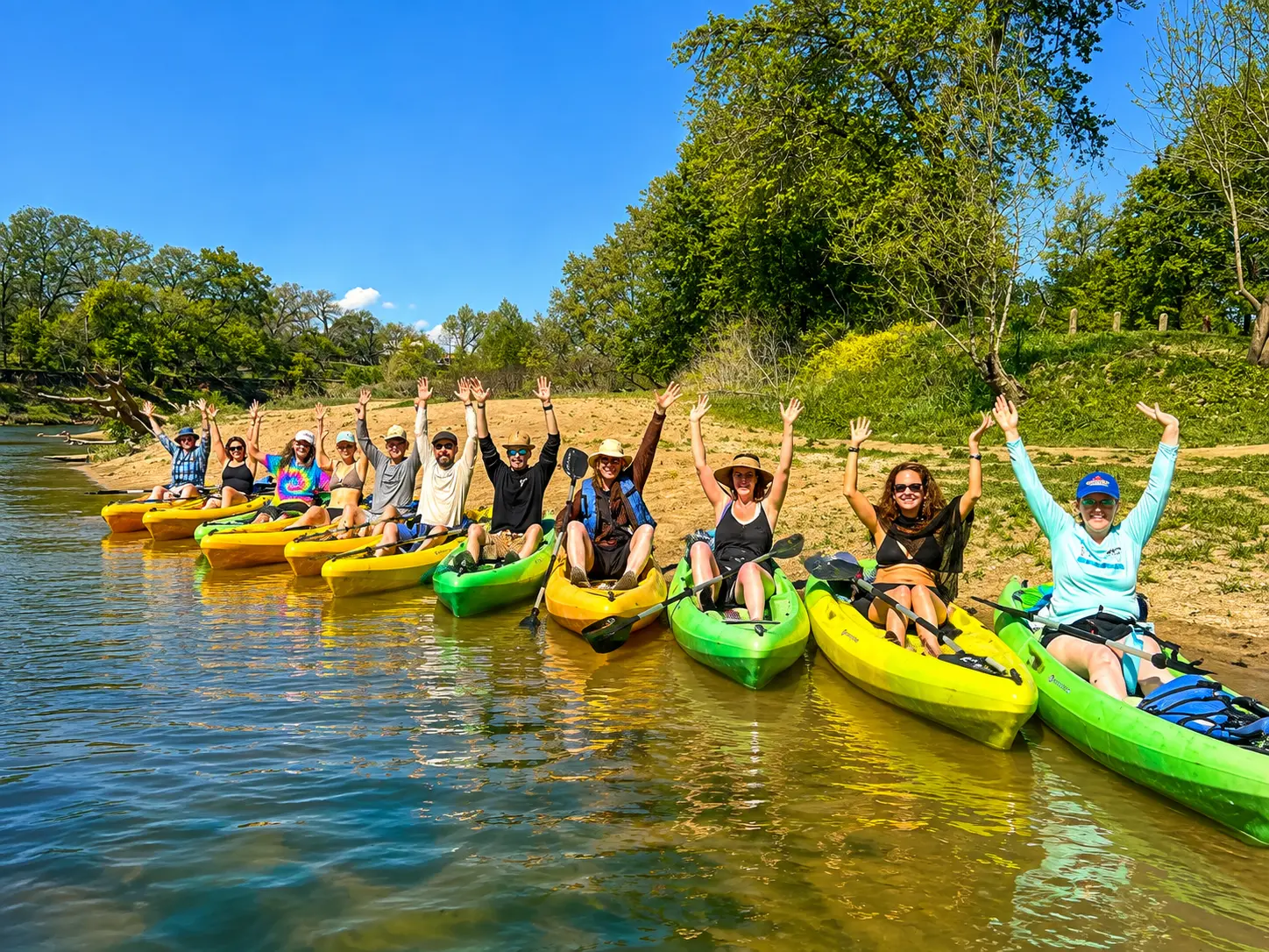 A large group of friends in colorful kayaks on the riverbank with their arms raised